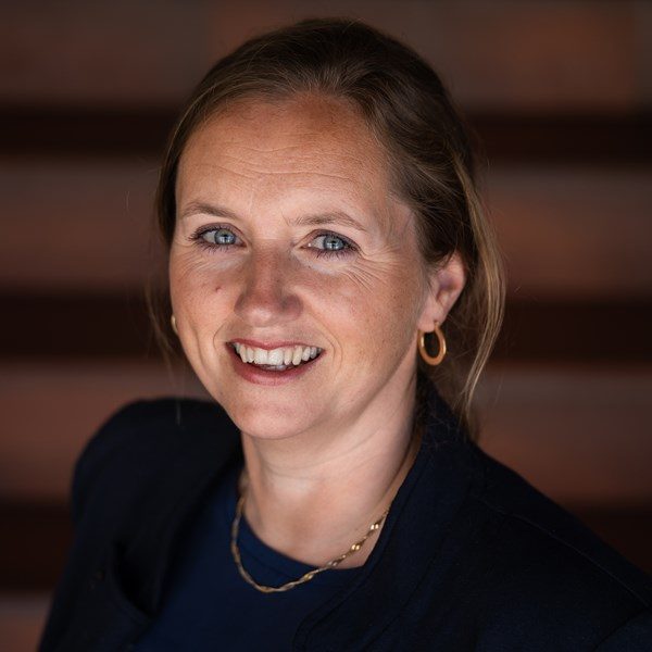 lise-beate-ringsby Picture of a woman from slightly above, smiling, with long hair a dark jacket and a necklace. The background is dark red and unsharp.